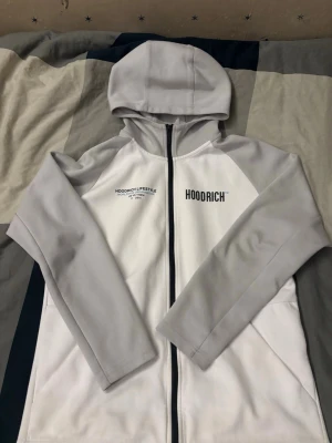 Vit och grå hoodie från Hoodrich - Snygg hoodie från Hoodrich i vitt med ljusgrå ärmar och huva. Dragkedja framtill och tryck med 'HOODRICH' på bröstet samt 'HOODRICH LIFESTYLE' och text på ryggen. Tillverkad i mjukt material, perfekt för en avslappnad streetwear-look.