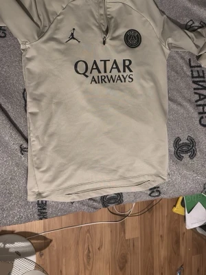 PSG tröja - Snygg beige tröja från Jordan x PSG med svart Paris Saint-Germain-märke och Jordan-logga på bröstet. Halvlång dragkedja framtill och Qatar Airways-tryck.