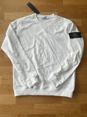 Vit sweatshirt från Stone Island - Säljer en stilren vit stickad tröja från Stone Island med klassisk rund hals och ribbade muddar. Tröjan har den ikoniska svarta patchen med gult och grönt Stone Island-märke på vänster ärm. Perfekt för dig som gillar streetwear och enkel design.  Helt ny och aldrig använd. Pris kan diskuteras