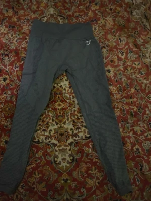 Grå seamless tights från Gymshark - Grå träningstights från Gymshark med hög midja och seamless design. Tightsen har diskreta mönster längs benen och en liten Gymshark-logga på höften. Perfekta för gymmet eller löpning. Liiitet hål som syns bild 2 o 3