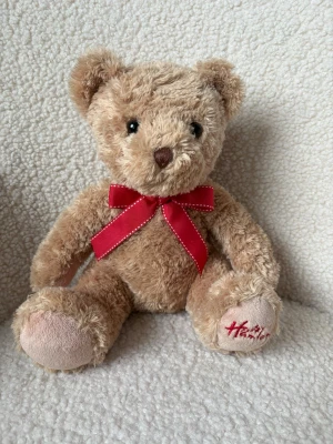 Mjuk nallebjörn med röd rosett - Söt och fluffig nallebjörn från Hamleys i ljusbrunt plyschmaterial. Nallen har en röd rosett runt halsen och broderad text på ena tassen. Perfekt som present eller dekoration.