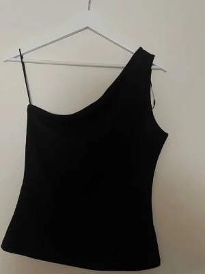 Svart one shoulder topp från H&M - Snygg svart one shoulder topp från H&M i stretchigt material. Toppen har en asymmetrisk design med en axel bar och en smal passform som framhäver siluetten. Perfekt att styla med jeans eller kjol för en trendig look.