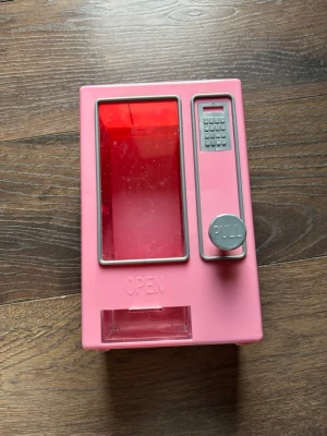 Rosa leksaksautomat i plast - Säljer en rosa leksaksautomat i plast med genomskinligt rött fönster och grå detaljer. Automaten har en knapp med texten 'PULL' och en liten lucka där man kan öppna och ta ut innehållet. Perfekt för småleksaker eller godis.