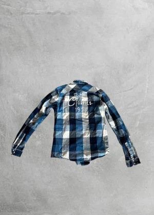 Blå och vit rutig Scotch & Soda skjorta - Snygg rutig skjorta i blått, vitt och grått med broderad text på ryggen. Skjortan har två bröstfickor med lock och knappar samt klassisk krage. Perfekt för dig som gillar en avslappnad men stilren look.