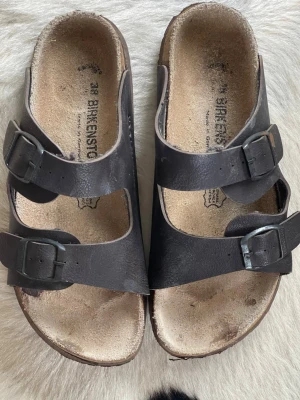 Bruna Birkenstock sandaler i storlek 38 - Klassiska bruna Birkenstock sandaler med två justerbara remmar och metallspännen i storlek 38. Fotbädden är i naturfärgat mocka och sulan är ergonomiskt formad för extra komfort. Perfekta för en avslappnad och trendig stil.