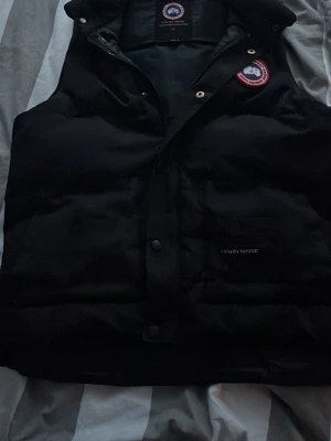 Canada Goose väst  - Snygg svart dunväst från Canada Goose med klassisk logotyp på bröstet och diskret broderad märkning. Västen har hög krage, tryckknappar och dragkedja framtill. Perfekt för lager-på-lager och håller dig varm under kyliga dagar.