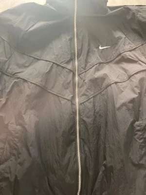 Nike vindjacka - Nike Windrunner | Svart | Stl XL En riktig streetwear-klassiker. Denna Nike Windrunner i svart nylon är både snygg och funktionell – perfekt för övergångssäsongen eller som ett stiligt lager ovanpå. Det ikoniska swoosh-logget och den karakteristiska paneldesignen gör den igenkännbar på direkten. Tidlös Nike-stil till ett kap av ett pris. ✔️ 