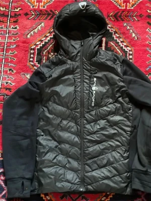 Svart vindjacka Sail Racing med huva - Snygg svart vindjacka från Sail Racing med huva och dragkedja framtill. Jackan har vadderad front och rygg i glansigt material, samt mjuka svarta ärmar. Logga på bröstet och sportig design. Perfekt för blåsiga dagar och outdoor-aktiviteter.