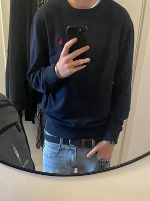 Mörkblå sweatshirt från Polo Ralph Lauren - Mörkblå sweatshirt från Polo Ralph Lauren med klassisk rund halsringning och röd broderad logga på bröstet. Tröjan har ribbade muddar vid ärmslut och nederkant. Perfekt för en clean och stilren look.