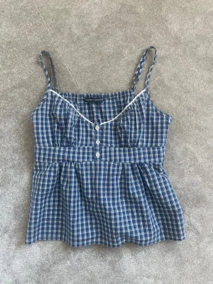 Blårutig linnetopp med spetsdetalj - Supersöt blårutig linnetopp med tunna axelband, knappar framtill och vit spets längs urringningen. Toppen har en markerad midja med knytband bak och lätt utställd nederdel. Perfekt för varma dagar och ger en chill vibe.