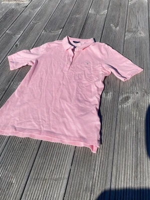 Ljusrosa pikétröja från Gant - Klassisk ljusrosa pikétröja från Gant med liten logga broderad på bröstet. Tröjan har krage, knappar framtill och korta ärmar. Tillverkad i mjuk bomull som känns skön mot huden. Perfekt för en avslappnad men ändå stilren look.