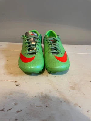 Nike Mercurial gröna fotbollsskor - Snygga gröna Nike Mercurial fotbollsskor med röd swoosh på sidan och framtill. Skorna har snörning och är tillverkade i ett lätt syntetmaterial som ger bra bollkontroll. Perfekta för dig som vill sticka ut på planen med stilrena och sportiga skor.