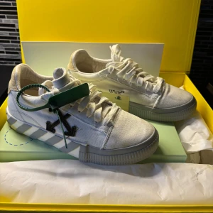 Off-White vita låga sneakers - Säljer ett par vita Off-White sneakers med ikoniska svarta pilar på sidan och randig sula i vitt och grått. Skorna har textdetaljerna 'THIS' och 'THAT' på hälen samt grönt tag. Tillverkade i canvas med klassisk snörning och chunky sula. Köpta på Mytheresa, kvitto och kartong finns kvar.