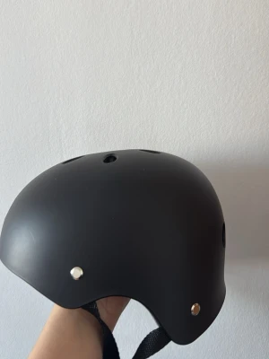 Svart skatehjälm med ventilationshål - Snygg svart skatehjälm/elsccoter/cykel med rundad form och flera ventilationshål upptill. Justerbart svart hakband med röd spänne för säker passform. Utsidan är matt och insidan har stötdämpande material. Perfekt för cykel, skateboard eller kickbike. Aldrig använd