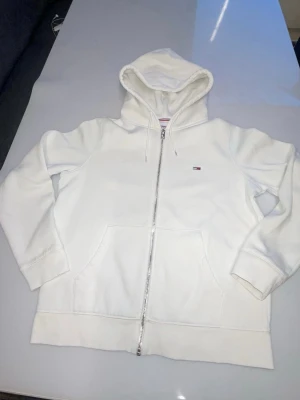 Vit kofta från Tommy Hilfiger - Vit kofta med dragkedja från Tommy Hilfiger. Klassisk design med huva och dragsko, två stora fickor framtill och liten broderad logga på bröstet. Tillverkad i mjuk bomull för en clean och enkel stil.