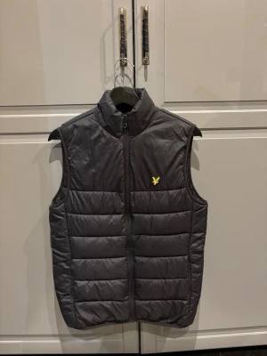 Svart dunväst från Lyle & Scott - Snygg svart dunväst från Lyle & Scott med gul logga på bröstet. Västen har hög krage, dragkedja framtill och quiltad design. Perfekt att slänga över en hoodie eller tröja för extra värme. Klassisk och stilren look som funkar till det mesta.
