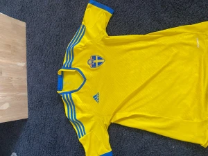 Svensk landslagströja Adidas gul/blå - Gul svensk landslagströja från Adidas med blå detaljer och SvFF-emblem på bröstet. Tröjan har klassiska blå ränder på axlarna och blå V-ringad krage. Perfekt för fotboll och supporterhäng. Hör av er vid frågor eller funderingar 