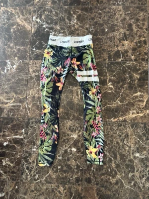Blommiga leggings från Stronger - Snygga leggings från Stronger med tropiskt blommönster i grönt, rosa, gult och vitt. De har bred resår i midjan med logga och vita ränder på ena benet. Tillverkade i stretchigt material som sitter skönt och följsamt.