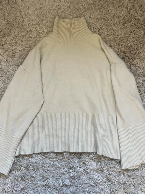 Beige polotröja med ribbade detaljer - Mysig beige stickad polotröja med bred ribbad kant vid ärmslut, nederkant och hals. Tröjan har en loose passform och långa ärmar, perfekt för kyliga dagar. Enkel och stilren design som passar till det mesta.