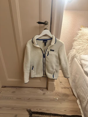 Vit fleecejacka från Patagonia XS - Säljer en vit fleecejacka från Patagonia i storlek XS. Jackan har blå detaljer vid dragkedjor och krage, tre fickor med dragkedja och mjukt, fluffigt material. Perfekt för lager på lager och riktigt skön att ha på sig under kyliga dagar.