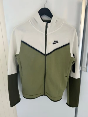 Nike hoodie i vitt och olivgrönt - Nike hoodie med dragkedja i vitt och olivgrönt. Snygg design med svart Nike-logga på bröstet och svarta detaljer längs bröstet. Huva och långa ärmar, samt fickor på sidorna. Tillverkad i mjukt och bekvämt material, perfekt för en avslappnad streetwear-look.OBS de finns en fläck på bröstet men den går nog bort i tvätten 