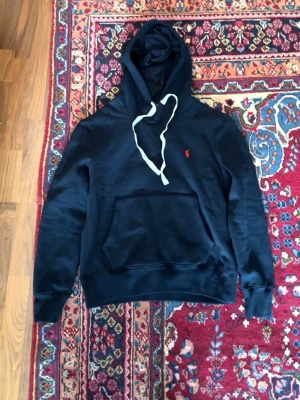 Mörkblå hoodie från Polo Ralph Lauren - Mörkblå hoodie från Polo Ralph Lauren med vit dragsko och klassisk känguruficka. Liten röd logga broderad på bröstet. Tillverkad i mjukt bomullsmaterial och har ribbade muddar vid ärmslut och nederkant. Perfekt för en avslappnad och stilren look.