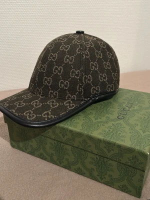 Ny G*cci brun keps med GG-mönster - Säljer en brun keps från G*vci med det klassiska GG-monogrammet över hela kepsen. Kepsen har böjd skärm med svart kant i läderimitation och är tillverkad i canvasmaterial. Perfekt för dig som vill ha en stilren och lyxig accessoar.