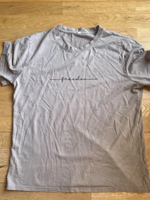 Beige t-shirt med texten freedom - Snygg beige t-shirt i bomull med svart broderad text 'freedom' på bröstet. Klassisk rund halsringning och korta ärmar. Perfekt basic-plagg för en avslappnad stil.