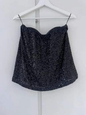 Svart glittrig bandeou topp från Gina Tricot - Säljer denna snygga fest topp från Gina täckt av glittrande paljetter. Toppen har en lätt utsvängd passform och mjukt innerfoder för extra komfort. Perfekt för dig som vill sticka ut med en glammig look.. Toppen har inga axelband.