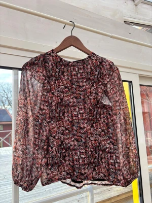 Blommig transparent blus med lång ärm - Säljer en blommig blus i transparent material med svart innertyg. Blusen har långa puffiga ärmar och ett mönster i rosa, vitt och svart. Rund halsringning och avslappnad passform. Perfekt för dig som gillar romantisk och trendig stil.