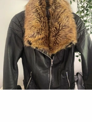 Svart skinnjacka med fuskpäls - Snygg svart skinnjacka med stor brun fuskpäls-krage och dragkedja framtill. Jackan har två snedställda fickor med dragkedja och coola sömdetaljer på axlarna. Perfekt för dig som vill ha en edgy look.