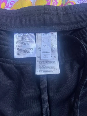 Svarta Adidas mjukisbyxor med vita ränder - Säljer ett par oanvända klassiska svarta mjukisbyxor från Adidas med de ikoniska tre vita ränderna längs benen och Adidas-logga på låret. Byxorna har dragsko i midjan och är tillverkade i ett mjukt, bekvämt material. Perfekta för chill eller träning.