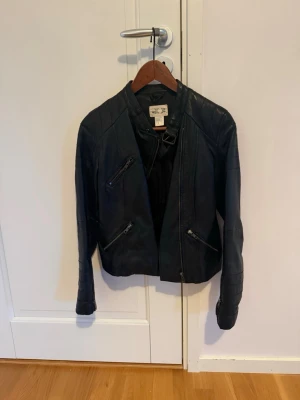 Svart skinnjacka med dragkedjor - Snygg svart skinnjacka från Ellos med coola detaljer som snedställda dragkedjor och ståkrage med spänne. Jackan har en figurnära passform och quiltade partier på axlar och ärmar. Perfekt för dig som vill ha en edgy look.