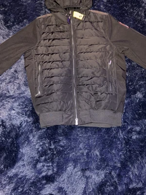 Parajumper jacka - Snygg svart pufferjacka från Parajumpers i storlek M. Jackan har huva, två dragkedjeförsedda fickor framtill och ribbade muddar vid ärmslut och nederkant. Tillverkad i lättviktigt material med quiltad front och släta ärmar. Perfekt för kalla dagar.