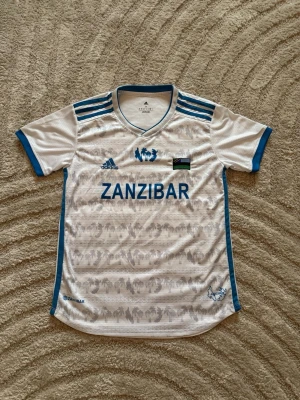 Vit Zanzibar fotbollströja Adidas - Snygg vit fotbollströja från Adidas med blå detaljer och texten 'ZANZIBAR' på bröstet. Tröjan har blå ränder på axlarna, blå kant vid ärmslut och hals, samt tryckta emblem och flagga. Tillverkad i lätt och ventilerande material, perfekt för träning eller match.