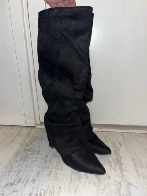 Svarta slouchy stövlar med klack - Säljer ett par svarta slouchy stövlar med spetsig tå och bred klack. Stövlarna har dragkedja på insidan och är tillverkade i ett mockaliknande material. Perfekta för att lyfta vilken outfit som helst med sin trendiga look.