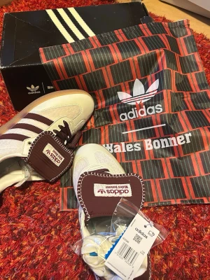 Adidas Wales Bonner sneakers vita/mörkröd  - Snygga Adidas Wales Bonner sneakers i vitt och mörkrött nästan brun  med klassiska ränder på sidan och detaljerad tunga. Skorna har mocka- och läderdetaljer, mörkröda nästan brun  skosnören och gummisula. Kommer med originalkartong och dustbag med Wales Bonner-logga. 42 i storlek men funkar i 41 och 43 lika bra    (SKORNA ÄR HELT OANVÄNDA)