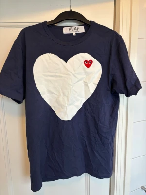 Blå Comme des Garçons PLAY t-shirt - Mörkblå t-shirt från Comme des Garçons PLAY med stort vitt hjärta och röd hjärtdetalj med ögon på bröstet. Klassisk rund hals och korta ärmar. Perfekt statement-plagg för dig som gillar streetwear och unika prints.