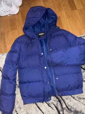 Blå pufferjacka från Urban Classics - Snygg blå pufferjacka från Urban Classics med huva och dragkedja. Jackan har tryckknappar framtill och är vadderad för extra värme. Lite smutsig vid ärmen men försvinner i tvätten! Perfekt för kalla dagar och har en klassisk, enkel design som passar till det mesta.