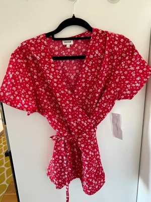 Röd blommig omlottblus från bubbelroom - Röd omlottklänning från bubbleroom med vitt blommönster och knytband i midjan. Blusen har v-ringning och korta ärmar, vilket ger en luftig och trendig look. Perfekt för sommaren och tillverkad i ett lätt material.