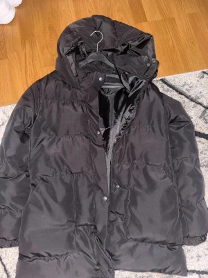 Svart jacka med huva - Säljer en svart jacka med huva i storlek S. Jackan har en puffig design, dragkedja och tryckknappar framtill samt två sidofickor. Jackan är lite längre, går ner till höfterna. Materialet är glansigt och jackan är perfekt för kalla vinterdagar. 