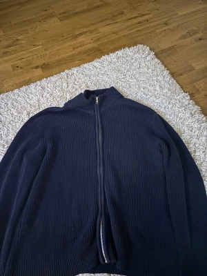 Mörkblå ribbad ziptröja - Säljer en mörkblå ribbstickad tröja med dragkedja framtill och hög krage. Tröjan har långa ärmar och är tillverkad i ett mjukt, tjockare material som passar perfekt när det är kyligt ute. Enkel och stilren design som funkar till det mesta.