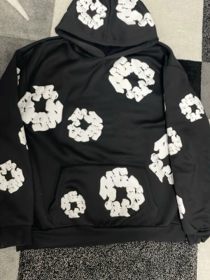 Denim Tears Hoodie Svart - Cool svart denim tears hoodie med huva. Hoodien har en klassisk känguruficka framtill och ribbade muddar vid ärmslut och nederkant. Perfekt för dig som gillar streetwear och unik design.