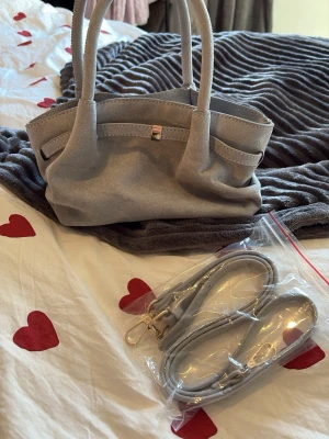 Ljusgrå handväska i mockaimitation - Trendig ljusgrå handväska i mjuk mockaimitation med dubbla handtag och avtagbar axelrem. Väskan har magnetlås och rymligt huvudfack med svart innerfoder. Perfekt accessoar för dig som gillar stilrena detaljer.