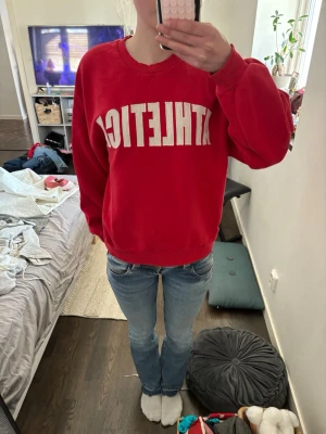 Röd sweatshirt med ATHLETICA-tryck - Cool röd sweatshirt med vit ATHLETICA-text framtill. Tröjan har en relaxed passform och rund halsringning. Perfekt för en avslappnad och sportig look. Tillverkad i mjukt material, troligen bomull eller polyester.