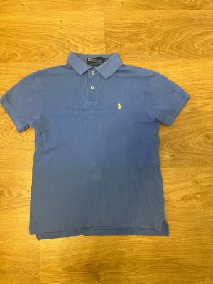 Blå piké från Polo Ralph Lauren - Klassisk blå pikétröja från Polo Ralph Lauren med gul broderad logga på bröstet. Slim fit-modell med korta ärmar, krage och knappar framtill. Perfekt för en clean och stilren look.