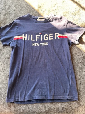 Blå Hilfiger Denim t-shirt - Snygg blå t-shirt från Hilfiger Denim med trycket 'HILFIGER NEW YORK' och röda samt vita detaljer över bröstet. Klassisk rund hals och korta ärmar. Tillverkad i mjuk bomull som känns skön mot huden. Perfekt för en avslappnad och trendig look. Nypris 349 Kr