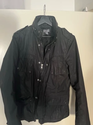 Svart vindjacka med dragkedja - Svart vindjacka med hög krage och flera fickor framtill. Jackan har dragkedja och tryckknappar, samt axelklaffar för en cool look. Perfekt för blåsiga dagar och enkel att matcha med olika outfits.