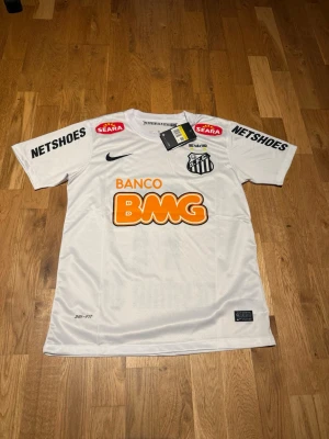 Santos 12-13 home kit Neymar Jr #11 - Säljer en vit Santos matchtröja från Nike med Neymar Jr och nummer 11 på ryggen. Tröjan har flera sponsorloggor, klubbmärke på bröstet och är tillverkad i lätt, andningsbart Dri-FIT-material. Perfekt för dig som älskar brasiliansk fotboll.