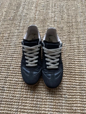 Svarta sneakers från Maison Margiela - Stilrena svarta sneakers från Maison Margiela med vita skosnören och beige sula. Ovandelen är i en mix av mocka och läder, med klassisk låg silhuett och diskreta detaljer. Perfekta för dig som gillar minimalistisk och tidlös design. Det är ett litet hål i hälen 